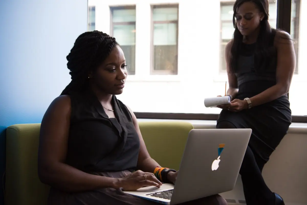 A young woman working at a laptop while a colleague reviews notes beside her